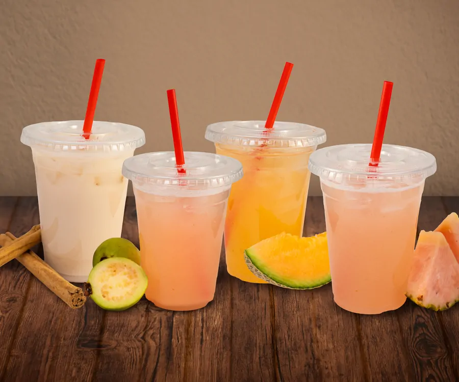 Four lidded clear cups of agua fresca with red straws on a wooden table: horchata beside cinnamon sticks, two pink ones, and a cantaloupe one, with cut guava, melon and watermelon around them.