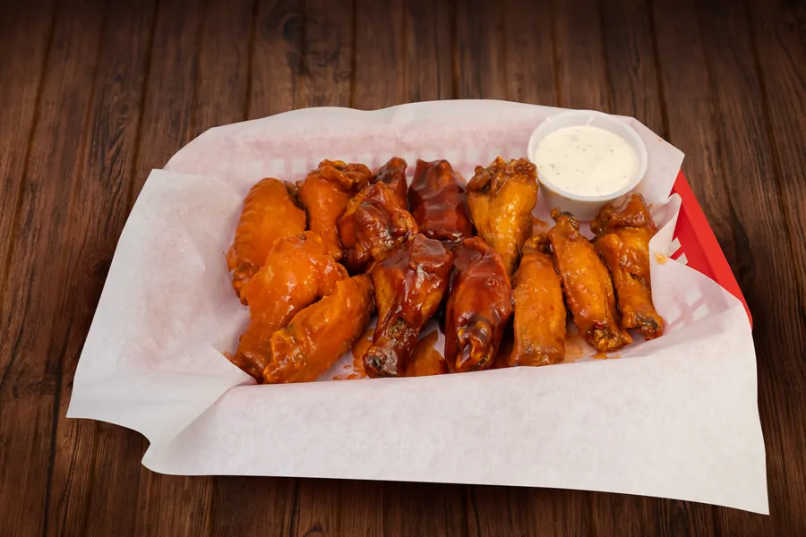 A paper lined basket of wings glazed in orange and dark red sauce, with a cup of white dipping sauce.