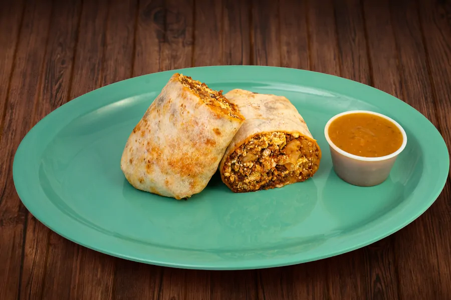 A breakfast burrito cut in half on a teal plate, the cut face showing egg, potato and meat, with a cup of sauce on the side.