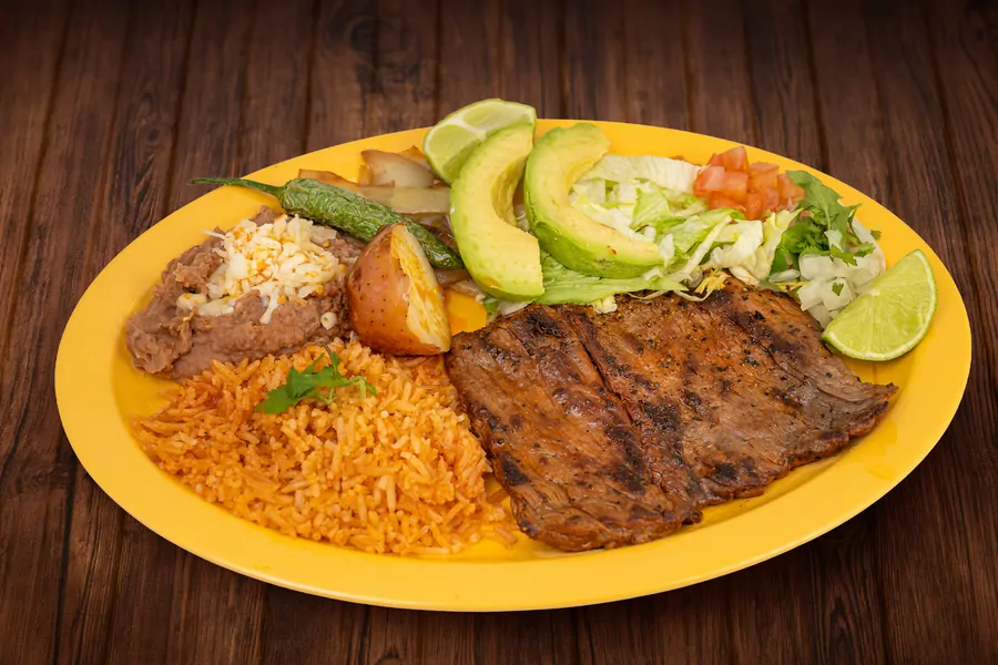 A grilled steak on a yellow plate with Mexican rice, refried beans topped with cheese, sliced avocado, lettuce, tomato, a grilled chile, a roasted potato and a lime wedge.