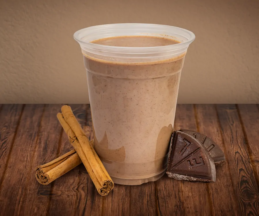A clear cup of champurrado, thick and light brown, on a wooden table with cinnamon sticks and wedges of Mexican chocolate beside it.