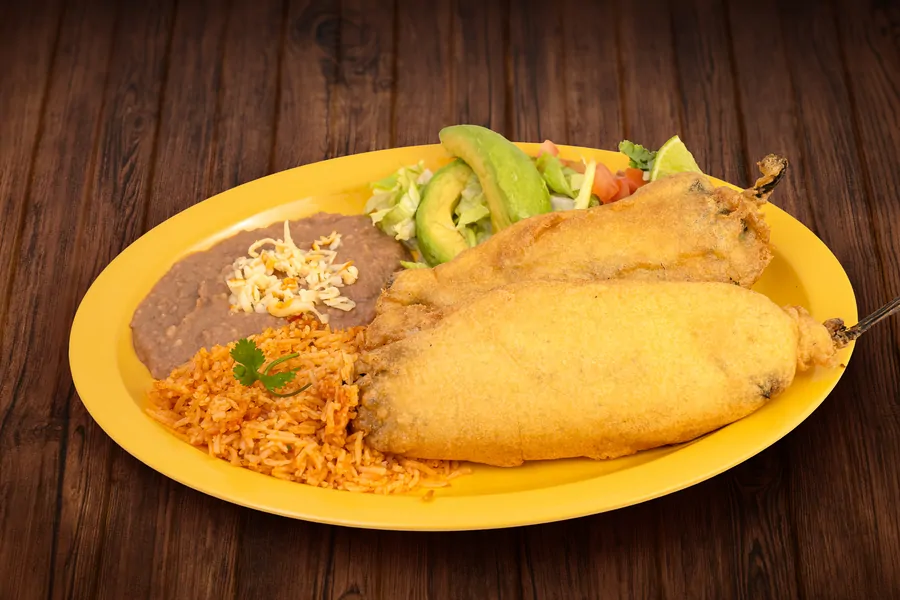 Two battered and fried chiles rellenos on a yellow plate with Mexican rice, refried beans topped with cheese, lettuce, avocado and tomato.
