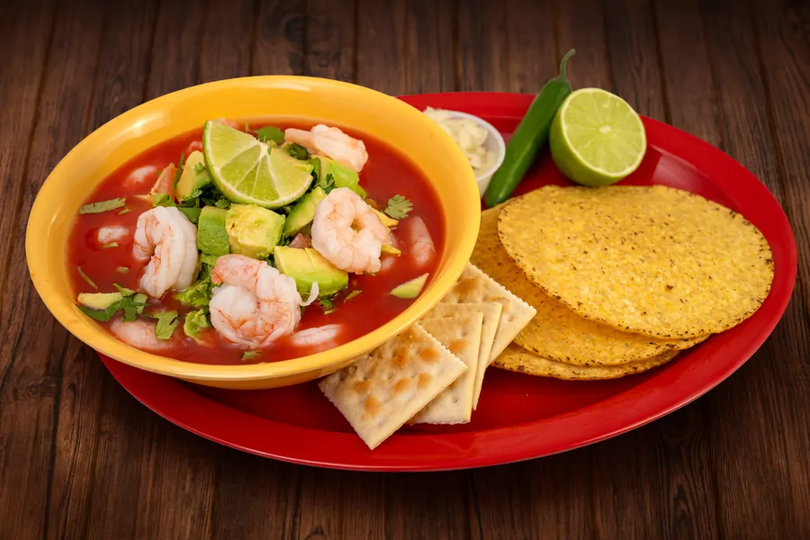 A yellow bowl of shrimp cocktail in tomato broth with avocado, cilantro and lime, on a red plate with tostadas, saltine crackers, a green chile and half a lime.