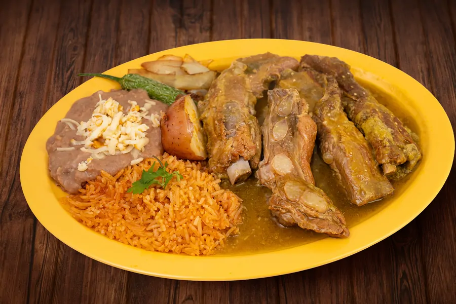 Pork ribs in sauce on a yellow plate with Mexican rice, refried beans topped with cheese, a roasted potato and a grilled chile.