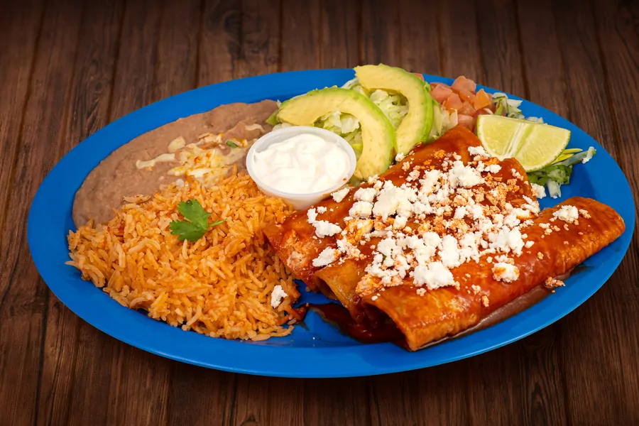 Enchiladas under red sauce and crumbled cheese on a blue plate with Mexican rice, refried beans, a cup of crema, avocado, lettuce, tomato and lime.