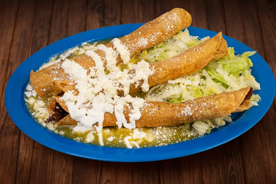 Four rolled and fried flautas on a blue plate over green salsa, topped with crema and crumbled cheese, with shredded lettuce.