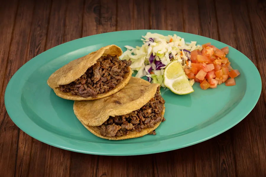 Two folded masa gorditas filled with chopped meat on a teal plate, with shredded cabbage slaw, diced tomato and a lime wedge.