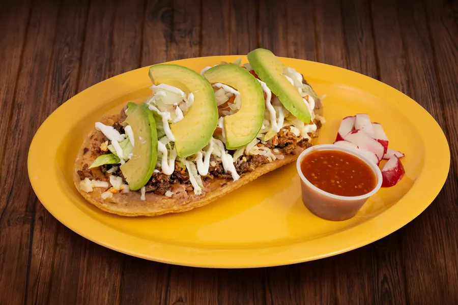 An oval masa huarache on a yellow plate layered with beans, chopped meat, lettuce, crema, cheese and sliced avocado, with a cup of red salsa and sliced radish.