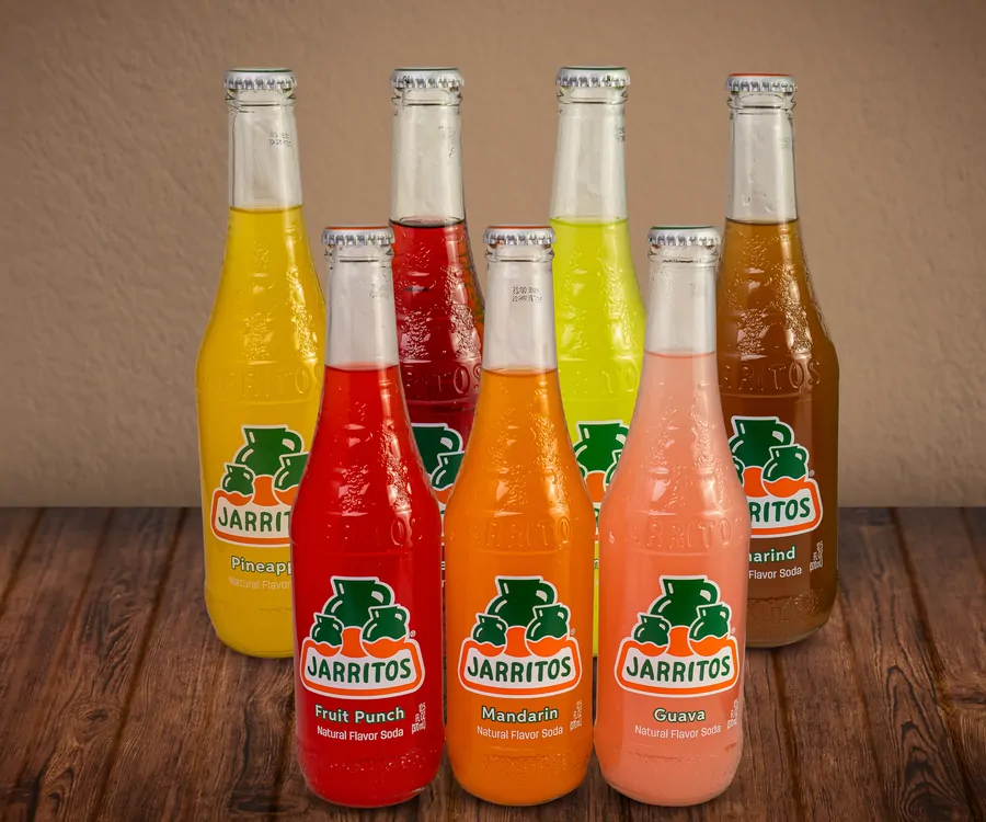 Seven glass bottles of Jarritos in two rows on a wooden table: pineapple, lime and tamarind behind, fruit punch, mandarin and guava in front.