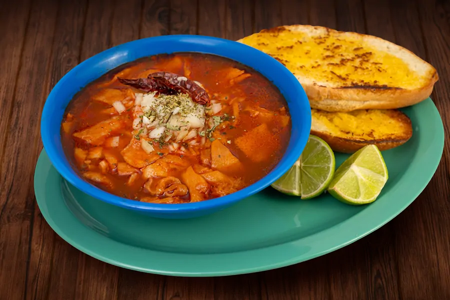 A blue bowl of menudo in red broth topped with chopped onion, oregano and a dried chile, with two pieces of grilled bread and lime wedges.