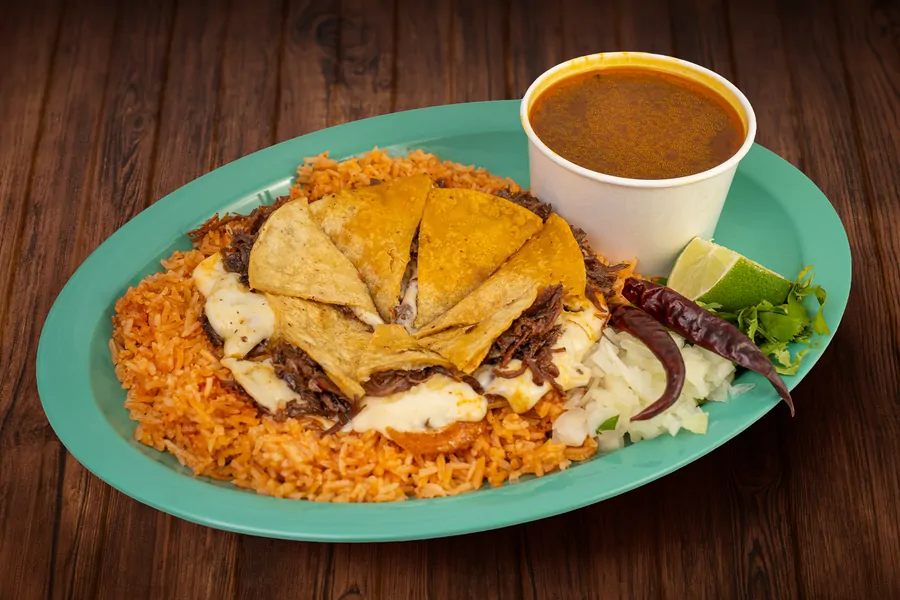 Quesabirria cut into wedges over a ring of Mexican rice on a teal plate, with a cup of consommé, a dried chile, onion, cilantro and lime.