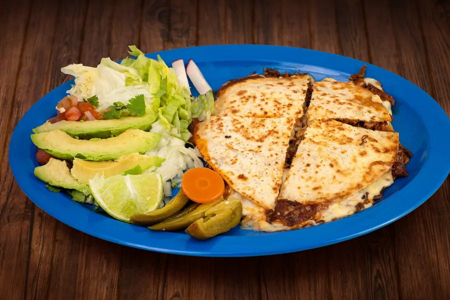 A large flour quesadilla cut in four on a blue plate with cheese browned at the edges, served with lettuce, avocado, pico de gallo, pickled jalapeños and lime.