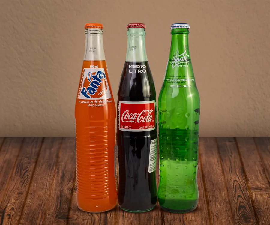 Three half litre Mexican glass bottles on a wooden table: Fanta orange, Coca-Cola and Sprite.