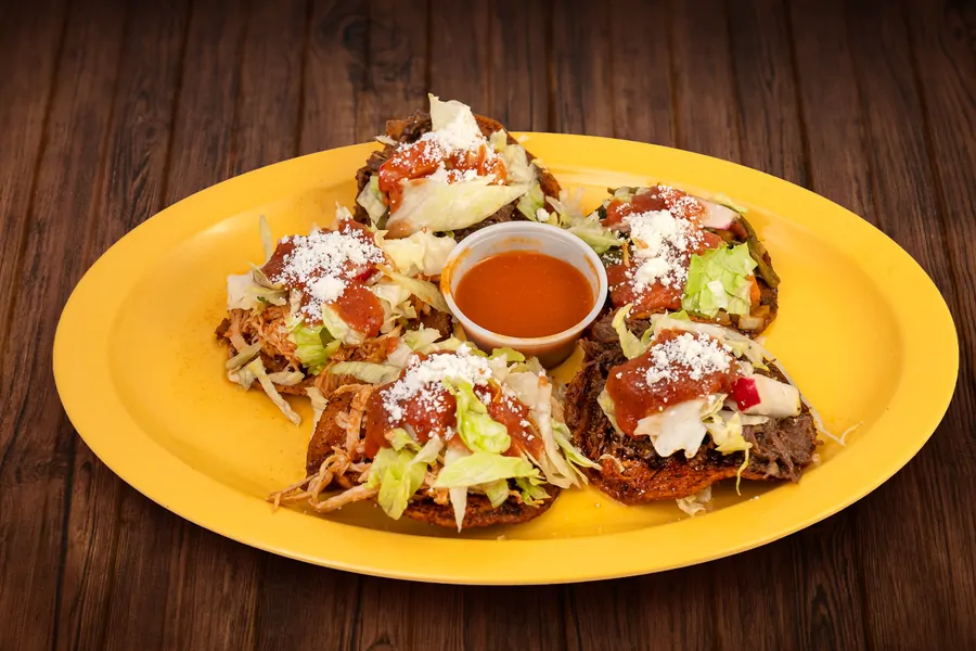 Five masa sopes on a yellow plate topped with different meats, shredded lettuce, red salsa and crumbled cheese, around a cup of red salsa.