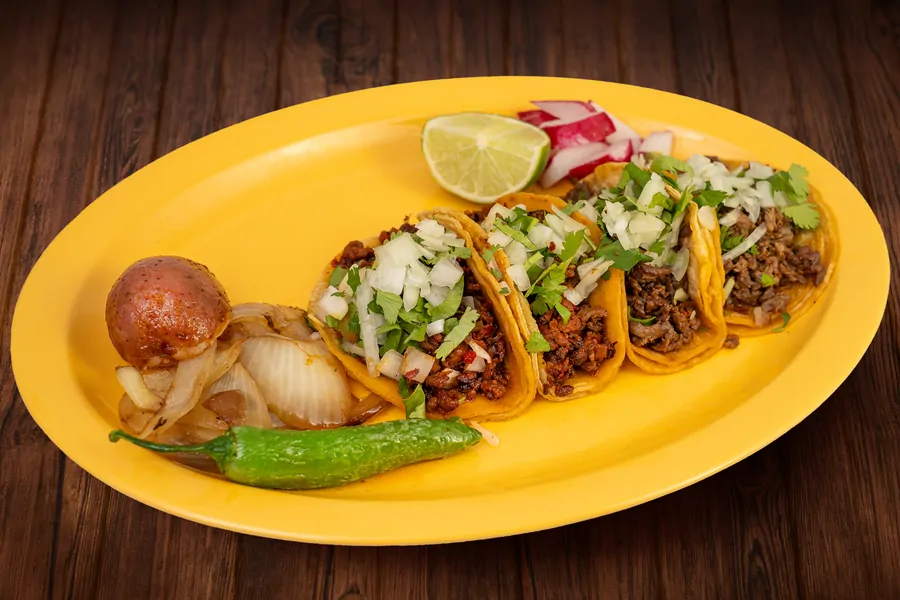 Four corn tortilla tacos on a yellow oval plate topped with chopped onion and cilantro, with grilled onion, a roasted potato, a whole green chile, lime and radish alongside.