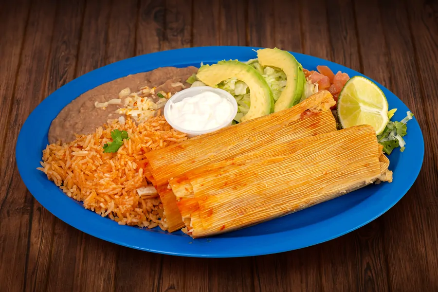 Two tamales in corn husks on a blue plate with Mexican rice, refried beans topped with cheese, a cup of crema, avocado, lettuce, tomato and lime.