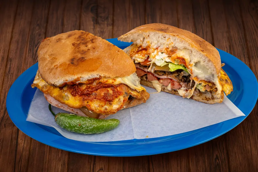 A torta cut in half on a blue plate on paper, layered with egg, ham, melted cheese, lettuce and tomato, with a whole green chile.