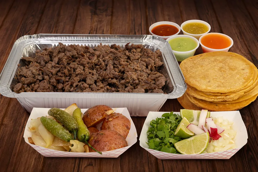 A foil half pan of chopped beef on a wood table with four cups of salsa, a stack of corn tortillas, a tray of grilled onion, jalapeños and roasted potatoes, and a tray of cilantro, lime, radish and diced onion.