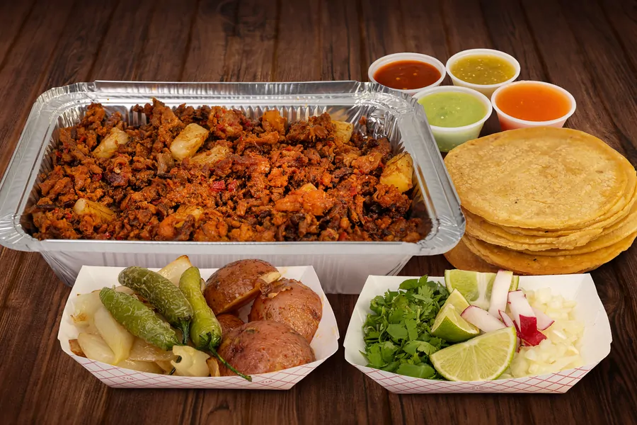 A foil half pan of red marinated meat with pineapple, with four cups of salsa, a stack of corn tortillas, a tray of grilled onion, jalapeños and roasted potatoes, and a tray of cilantro, lime, radish and diced onion.