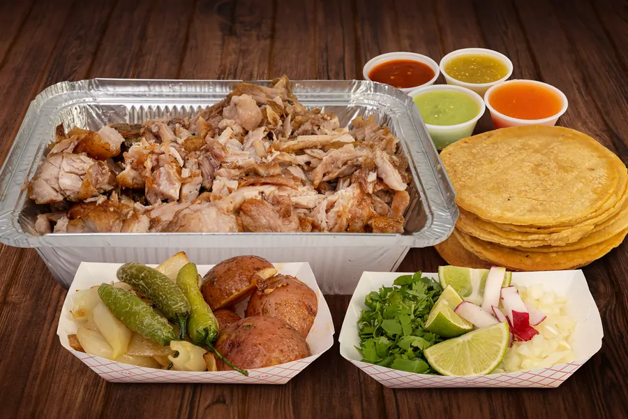 A foil half pan of shredded pork, with four cups of salsa, a stack of corn tortillas, a tray of grilled onion, jalapeños and roasted potatoes, and a tray of cilantro, lime, radish and diced onion.