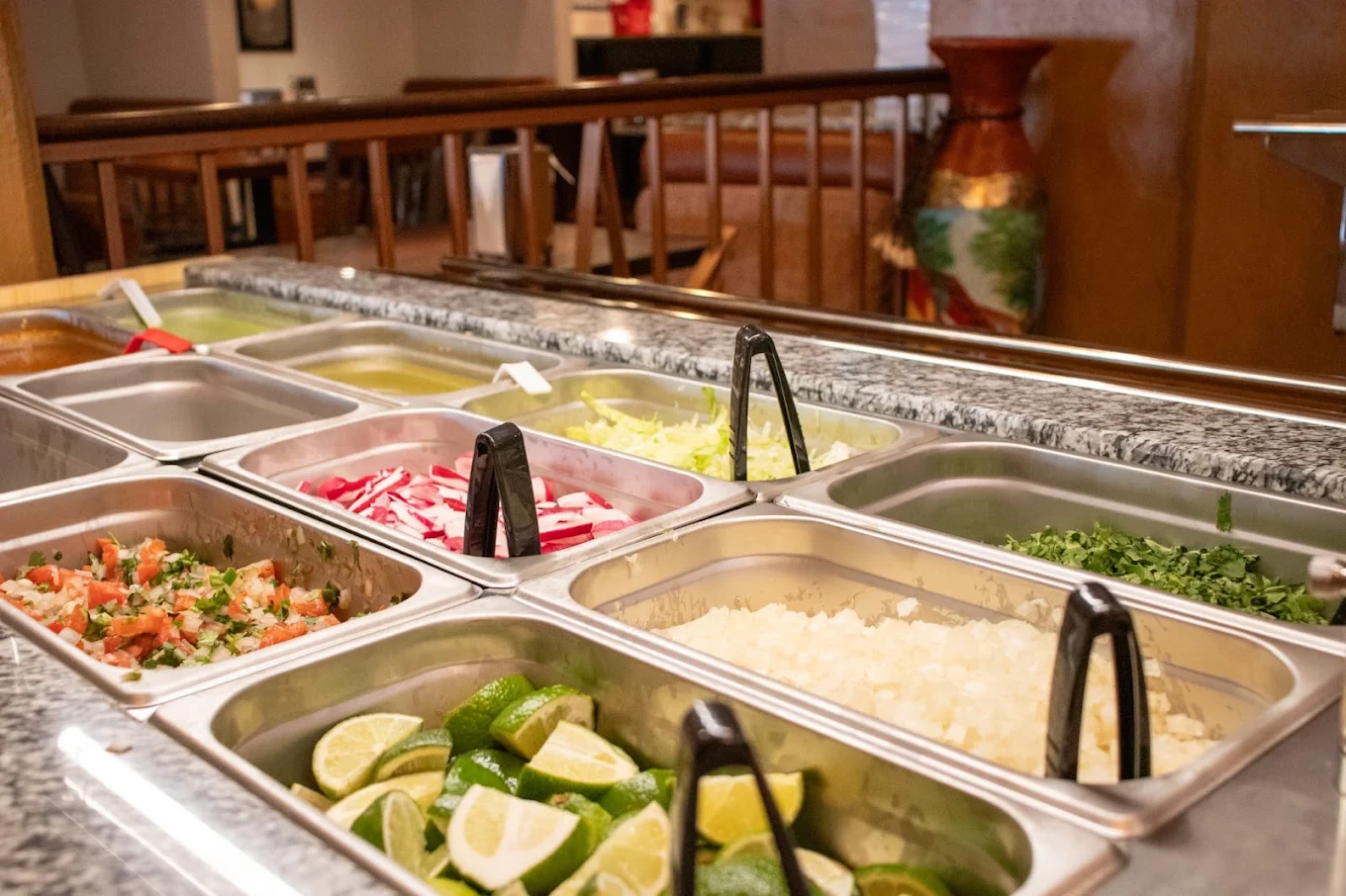 The salsa bar, with trays of pico de gallo, pickled radish, chopped onion and cilantro, grilled serrano peppers and lime wedges under warm light.
